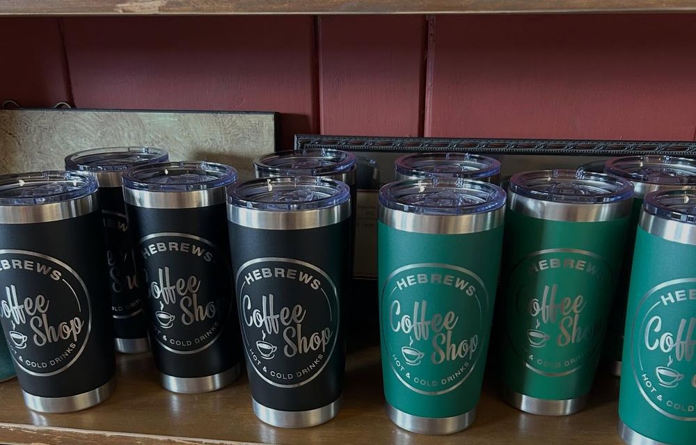 Black and green insulated Hebrews tumblers next to bags of roasted beans